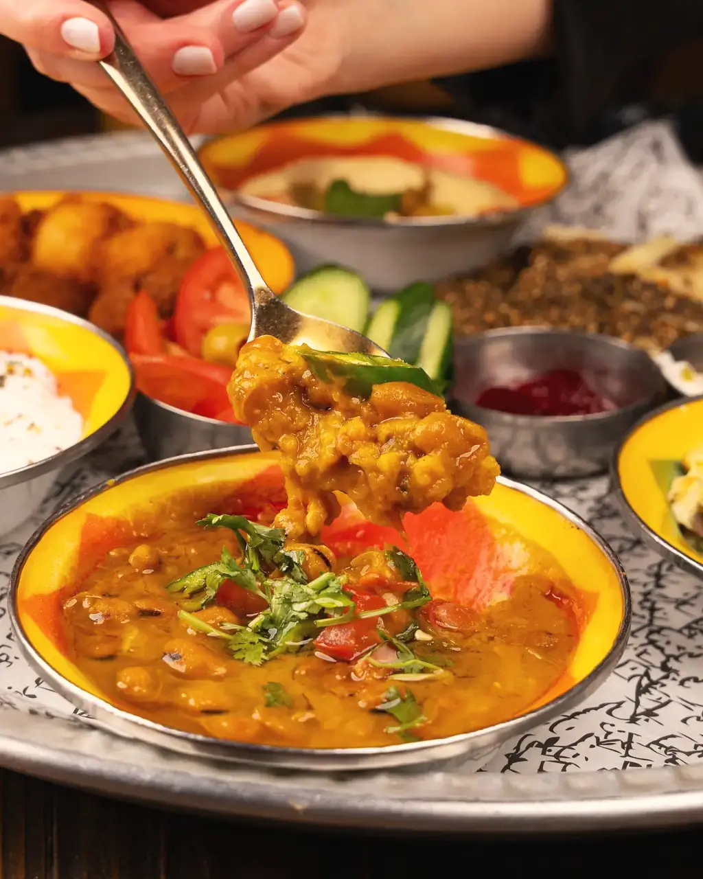 A spoon lifting dal from a steel bowl on a thali with rice, sabzi and salad